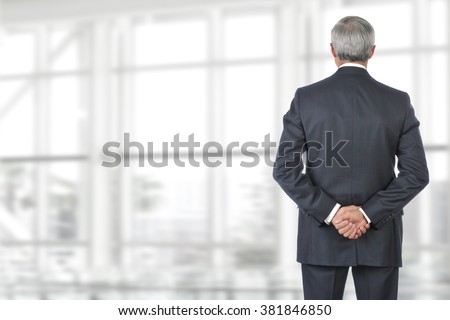 Portrait of smiling senior businessman standing looking out of office window with his hand behind his back. Royalty-Free Stock Photo #381846850