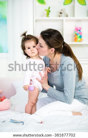 Young mom kissing her baby girl on the bed.Baby standing and looking at camera.Shallow doff