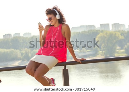 Young woman siting on fence and listening music using a smart phone outdoors.