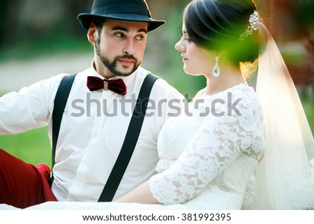 Stylish bride and groom sit on grass in rays of setting sunlight. Man in hat with bow tie and suspenders, red pants languidly looks at a woman with professional hair styling. A couple of newlyweds.