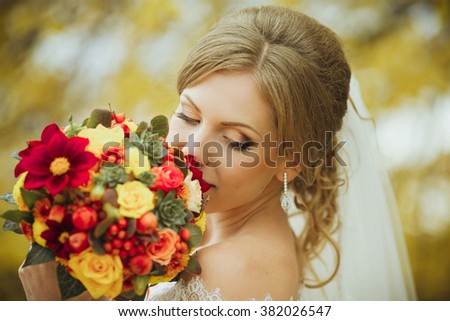A happy blonde bride holding a wedding bouquet with autumn trees on a background.