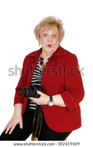 A very surprised looking senior woman sitting in a red jacket, holding a
camera in her hand, isolated for white background.
