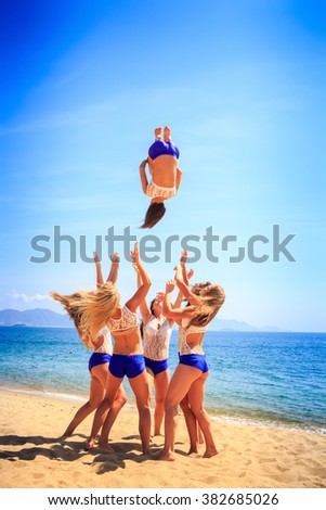 squad of six cute cheerleaders in white blue uniform performs Back Tuck Basket Toss on beach against azure sea