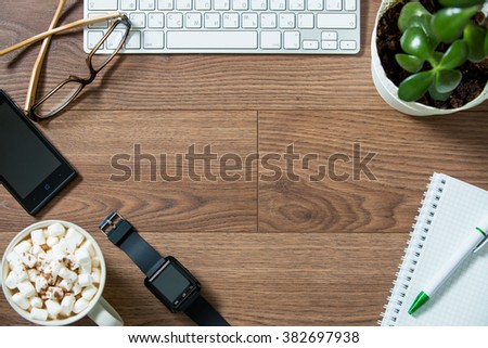 Business workplace from top, flay lay. Keyboard, smart watch, smart phone, glasses, paper notebook, hot chocolate with marshmallow on the dark wooden desk