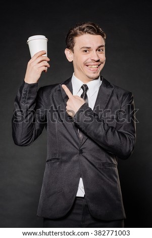young businessman in black suit holding a white blank paper cup of coffee with a white plastic cap . emotions, facial expressions, feelings, body language, signs. image on a black studio background.