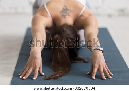 Beautiful young woman with flower tattoo on her back working out indoors, doing forward bend yoga exercise on blue mat, Balasana, Child Pose, Ardha-Kurmasana (Half Tortoise Pose), close-up Royalty-Free Stock Photo #382870504