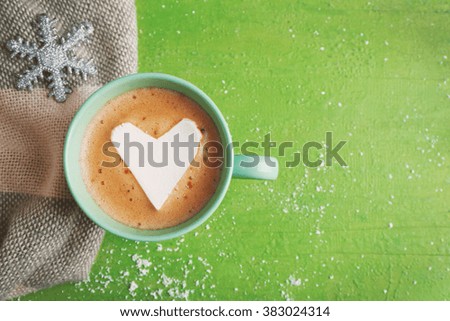 Cup of hot cappuccino with heart marshmallow, snowflake and warm scarf on green background, close up