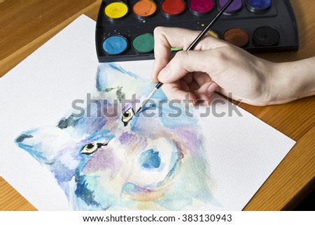  The artist paints a picture using brush and watercolor colorful painting set.  Hand with brush in process drawing picture Wolf. Wooden background. Modern art. Leisure. Artistic retouching.