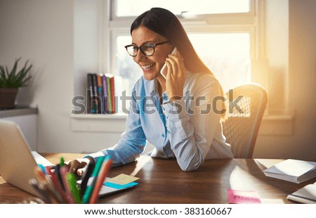 Business woman working on laptop smiling talking on phone sun coming through window Royalty-Free Stock Photo #383160667