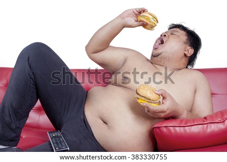 Picture of a young obese man watching tv while sitting on the sofa and enjoy two burger, isolated on white background