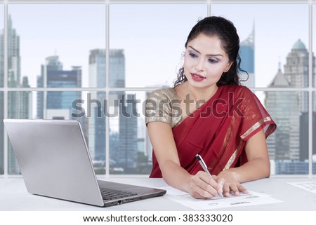 Image of pretty indian woman working with laptop and writing document in the office while wearing traditional clothes
