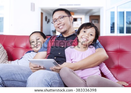 Picture of two happy children and their father sitting on the couch and smiling at the camera while holding a digital tablet at home