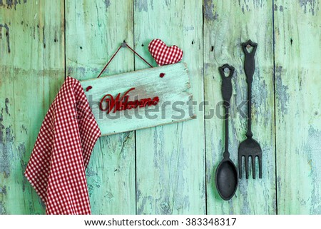 Welcome sign with red checkered gingham linen and heart hanging on antique rustic mint green wood background by black cast iron spoon and fork; Valentine's Day and love concept background