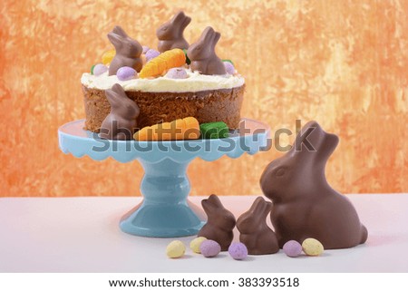 Easter Carrot Cake decorated with mini fondant carrots and chocolate bunnies on a white wood table with orange background. 