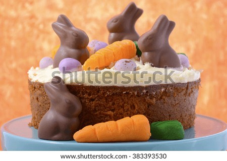 Easter Carrot Cake decorated with mini fondant carrots and chocolate bunnies on a white wood table with orange background, closeup.  