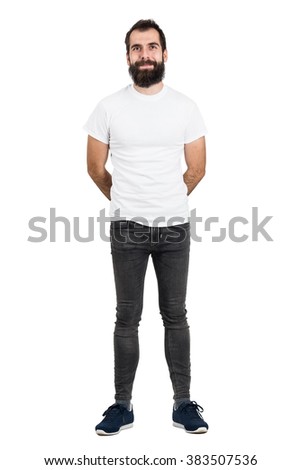 Happy bearded man with hands behind back in white t-shirt and tight jeans looking at camera. Full body length portrait isolated over white studio background.  Royalty-Free Stock Photo #383507536