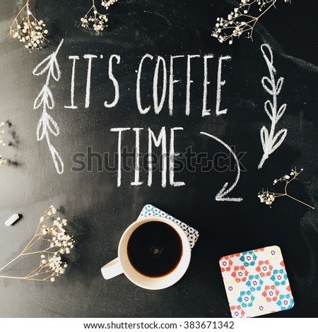 Phrase "It's coffee time" written in calligraphy style on black chalkboard with white flowers and a cup of coffee. Flat lay