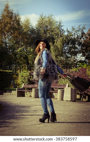 Beauty fashionable young woman having fun in park. Model wearing modern clothes in motion playing with her black bag outdoor.
