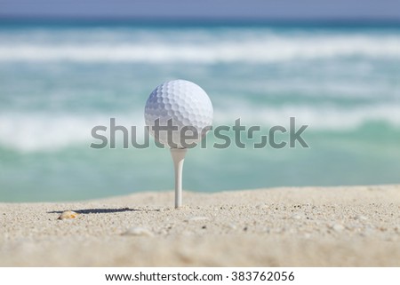 White golf ball on tee in sand of beach with soft focus ocean waves behind