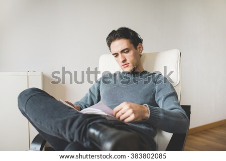 Young handsome caucasian man sitting on the armchair in his house, looking downward reading a book - student, knowledge, culture concept