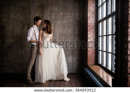 couple embrace on a background of textured walls in the loft, the bride in a wedding dress and beige with a bouquet of anemones in hand, groom in beige trousers and a white shirt with suspenders Royalty-Free Stock Photo #384093922