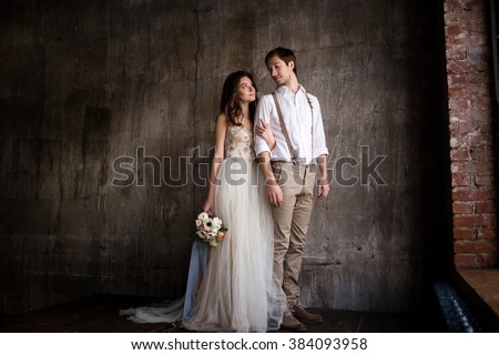 couple embrace on a background of textured walls in the loft, the bride in a wedding dress and beige with a bouquet of anemones in hand, groom in beige trousers and a white shirt with suspenders Royalty-Free Stock Photo #384093958