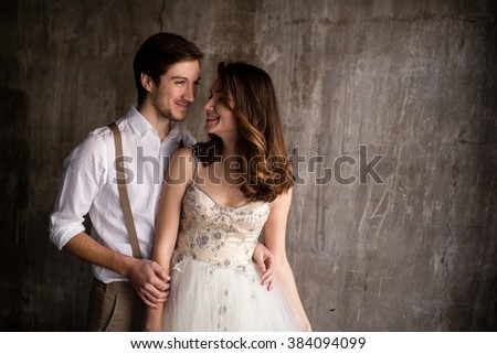 couple embrace on a background of textured walls in the loft, the bride in a wedding dress and beige with a bouquet of anemones in hand, groom in beige trousers and a white shirt with suspenders Royalty-Free Stock Photo #384094099