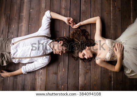 couple lying on the wooden floor and touching her hands, the bride in beige pants with suspenders and a white shirt, the bride in a beige dress Royalty-Free Stock Photo #384094108