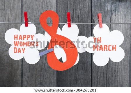 Happy Women's Day greeting message on white flower hanging on red pegs on a line against wooden background for International Womens Day, March 8.