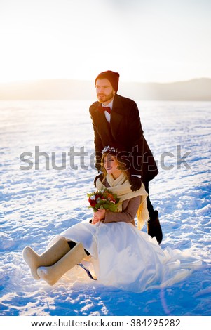 the groom rides the bride on the sledge on the stiffened lake against mountains. The bride in hands has a bouquet of flowers