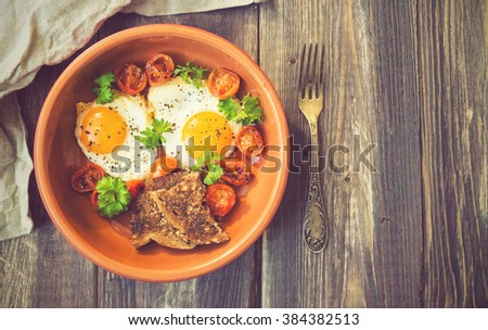 Fried eggs with cherry tomatoes and parsley in clay dish on rustic wooden background. Vintage toned picture.