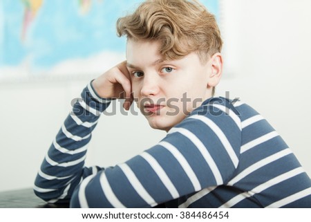 Head and Shoulders Portrait of Boy with Blond Wavy Hair Sitting at Desk with Head Resting on Hand in Classroom with World Map in Background