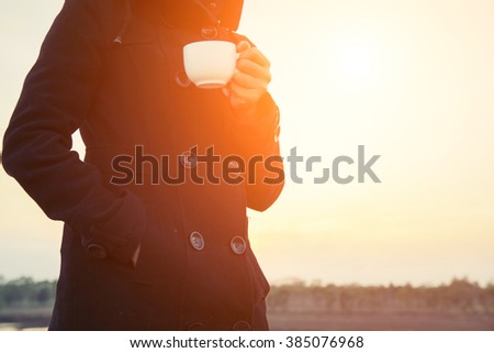 young woman's hands holding a cup of coffee, wearing black coat in the morning