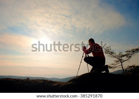 Nature photographer with tripod on cliff and thinking. Dreamy fogy landscape, orange misty sunrise in a beautiful valley below