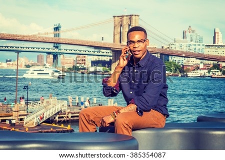 African American Man traveling, working in New York. Wearing blue shirt, brown pants, glasses, a college student sitting by river, talking on phone. Boat, Manhattan, Brooklyn bridges on background.
