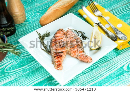 Grilled steak of salmon with rosemary and lemon in square plate  on  turquoise colored wooden table, Baguette, silver cutlery on yellow napkin at white polka dots, wooden saltcellar and pepper mill