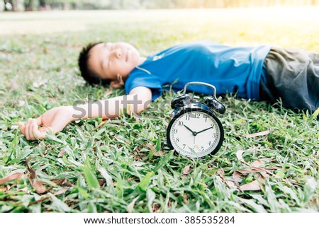 Vintage color tone, Selective focus on the classical black alarm clock model, in front of the sleeping young boy on green lawn in the park in day time.