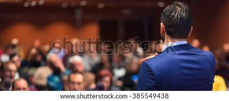 Rear view of speaker giving a talk on corporate Business Conference. Audience at the conference hall. Business and Entrepreneurship event. Panoramic composition. Royalty-Free Stock Photo #385549438