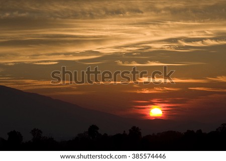 Crimson sunset, sun setting behind crest of mountain hill with clouds in sky reflecting orange and yellow