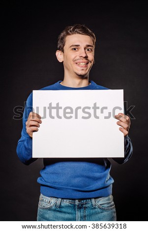 Happy young man holding white blank panel with space for text on black background.