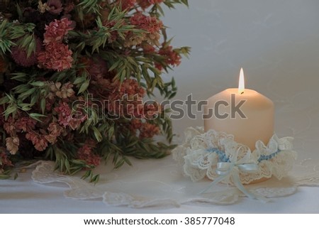 Single candle in a wedding garter with a bouquet of dried oats and flowers on festive embroidered white tablecloth