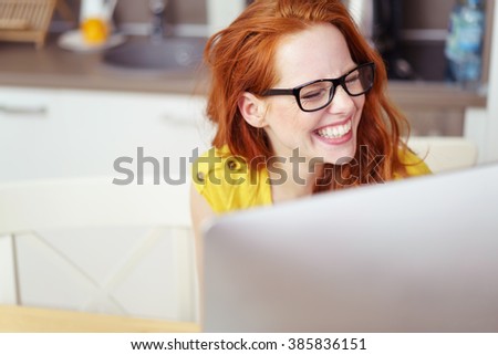 Head and Shoulders of Young Woman with Red Hair Wearing Eyeglasses and Laughing Joyfully While Working on Computer with Over-Sized Monitor at Home in Kitchen Royalty-Free Stock Photo #385836151