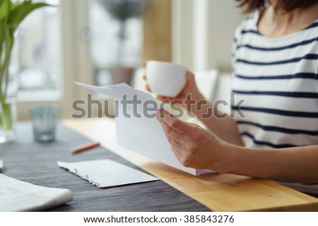 Woman reading a document at the dining table as she enjoys a morning cup of coffee, close up view of her hands