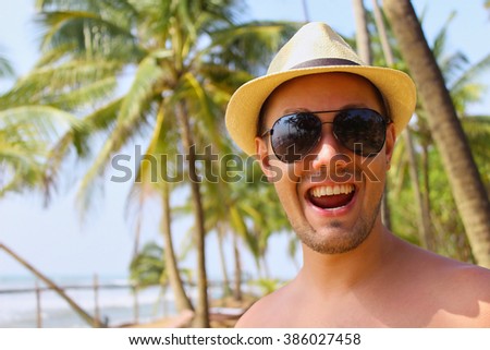 Young man smiling on a tropical  summer beach, happy mood, wearing sunglasses, palm trees background. Traveling vacation photo.