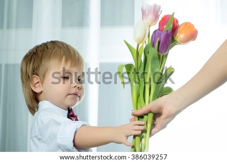 Happy smiling cute little boy gentleman with blonde hair in white shirt and bow tie giving beautiful fresh spring bouquet of colorful tulips for mothers day holiday, horizontal picture