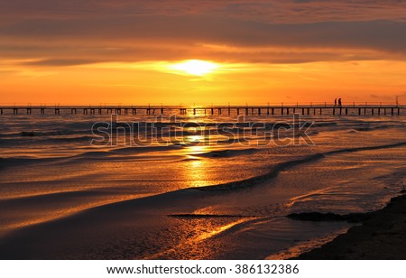 Golden natural sea sunset view of jetty or small bridge at horizon with people 
silhouette and orange sky landscape. Sunset or sunrise reflection in nature with sun in clouds above black sea scenery. 