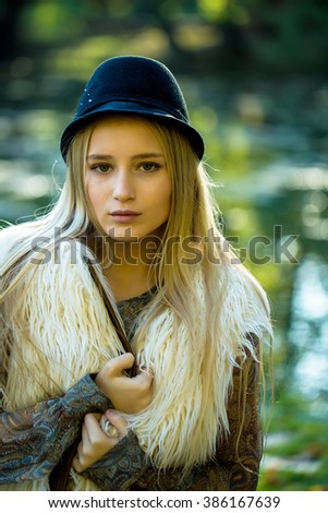 Portrait of young beautiful thoughtful woman with long blonde hair in stylish fur autumn waist coat and blue elegant hat outdoor on blurred natural background, vertical picture