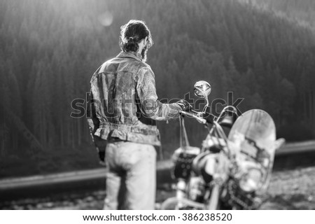 Long haired biker standing near his custom made cruiser motorcycle, wearing leather jacket and jeans. Looking into distance. Shot from the back. Black and white