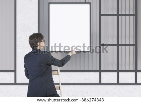 Businessman on ladder fixing blank frame on wall. Concept of office decoration.