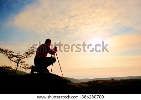 Nature photographer with tripod on cliff and thinking. Dreamy fogy landscape, orange misty sunrise in a beautiful valley below